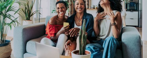 3 women sitting on a sofa talking, a laptop sits on a coffee table in front of them
