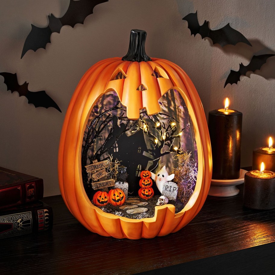 A large carved pumpkin displaying a lighted miniature Halloween scene inside featuring a haunted house, bare trees, a ghost, and smaller jack-o'-lanterns, sitting on a table next to lit black candles.