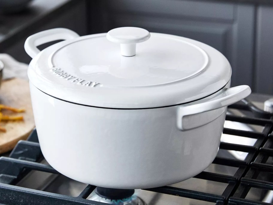 A white Bobby Flay 5.5-quart enameled cast iron Dutch oven with a matching lid and side handles, sitting on a stovetop burner.