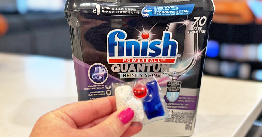 hand holding a dishwasher pod in front of a tub of Finish Quantum Infinity Shine tablets