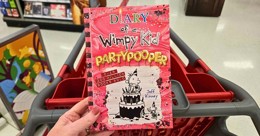 hand holding a Diary of a Wimpy Kid Partypooper book, Target cart behind it