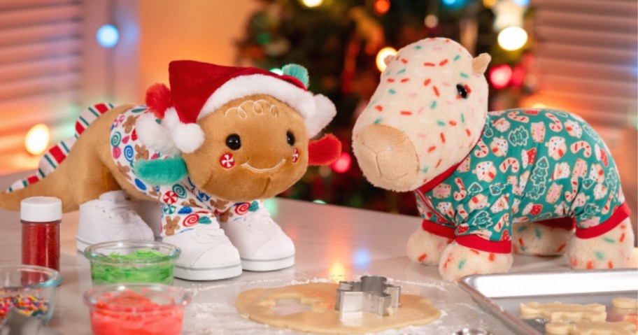 a plush gingerbread axolotl in a santa hat and a plush capybara in a holiday shirt on a table with Christmas decor