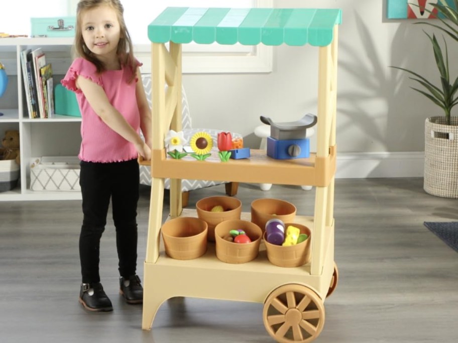 a little girl next to a toy farmstand with toy plants, flowers, and flower pots