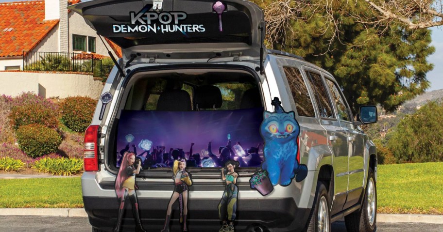 A silver SUV decorated for Halloween with a K-Pop Demon Hunters trunk-or-treat kit featuring character cutouts, a concert backdrop, and a blue cat illustration.
