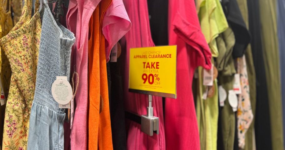 A yellow 90% off clearance sign in front of shirts and sun dresses