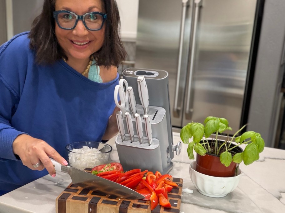 A woman in a blue shirt smiling while slicing red peppers and onions on a wooden cutting board next to a Ninja Foodi knife block and a potted basil plant.