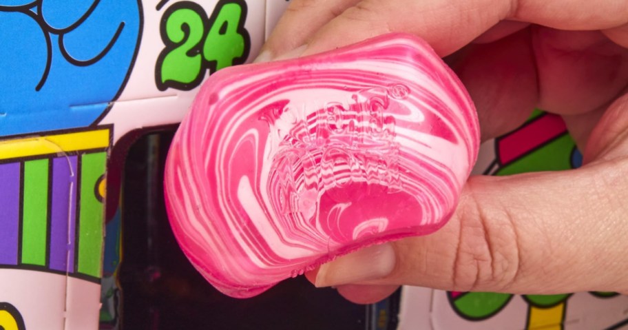 A hand holding a pink and white marbled NeeDoh fidget toy in front of a colorful advent calendar box.