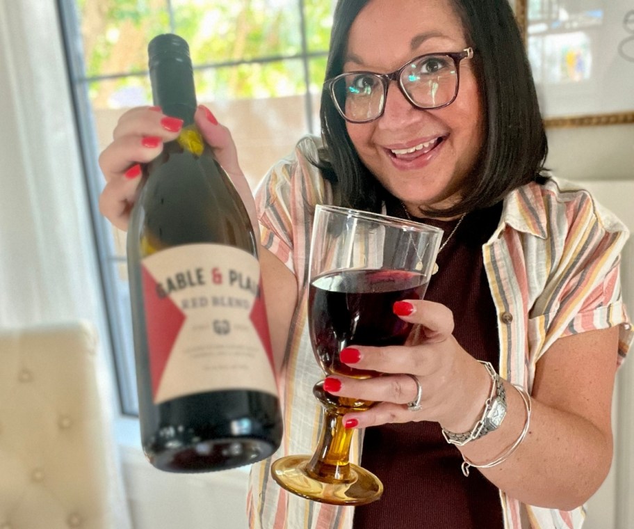 a woman holding a glass of wine and offering a wine bottles