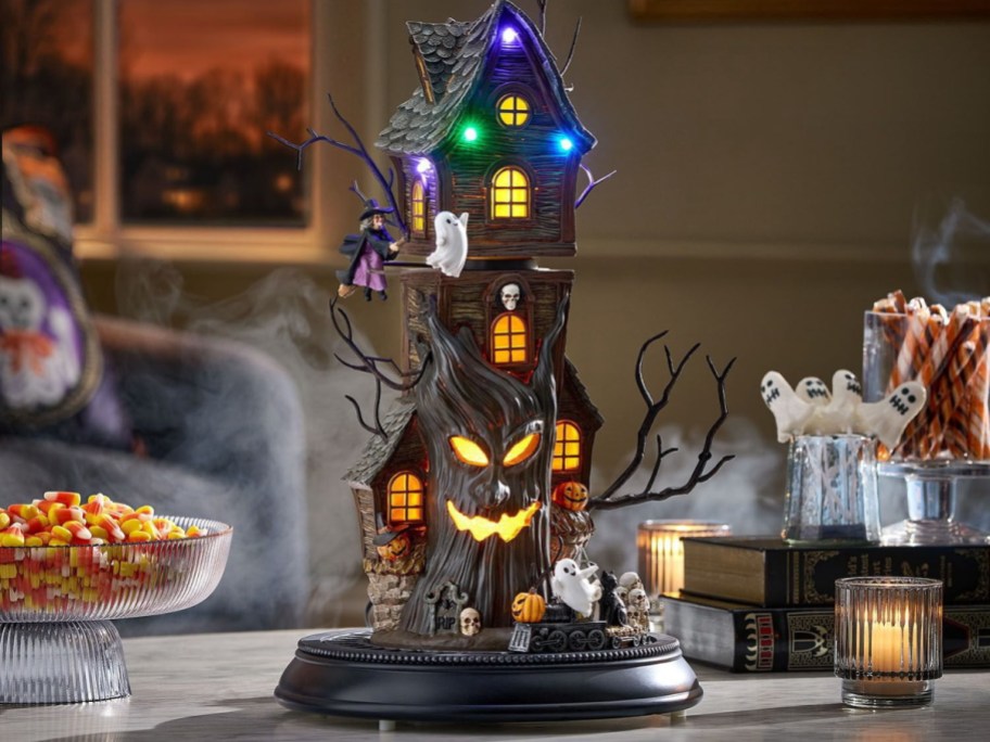 A lighted Halloween haunted house decoration featuring a spooky tree with a glowing carved face, lit windows, ghosts, a flying witch, and pumpkins, surrounded by fog.
