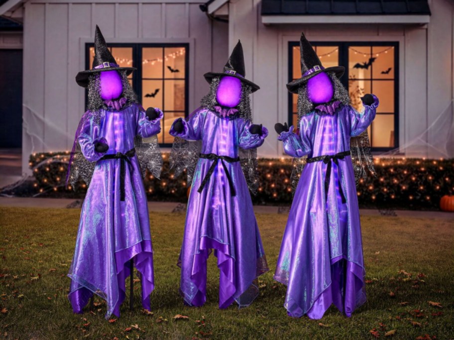Three lighted Halloween yard decorations of witches with glowing purple faces, wearing shiny purple dresses and black pointed hats, standing on a lawn in front of a house.