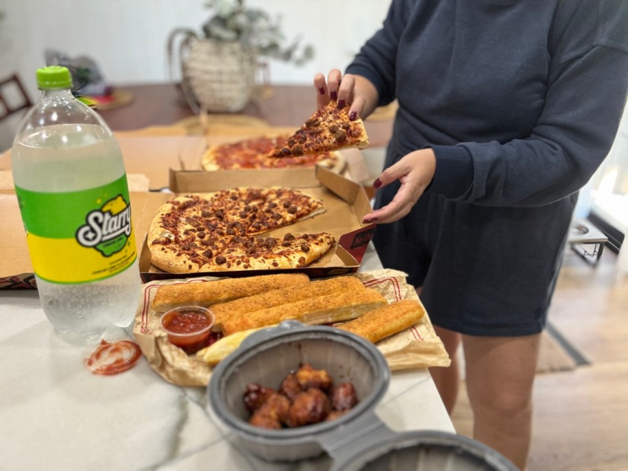 woman lifting a slice of pizza out of a box with wings, breadsticks and another pizza in the background