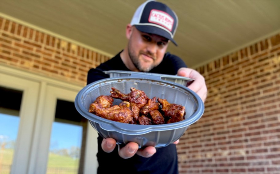 smiling man holding a container of chicken wings extended towards the camera