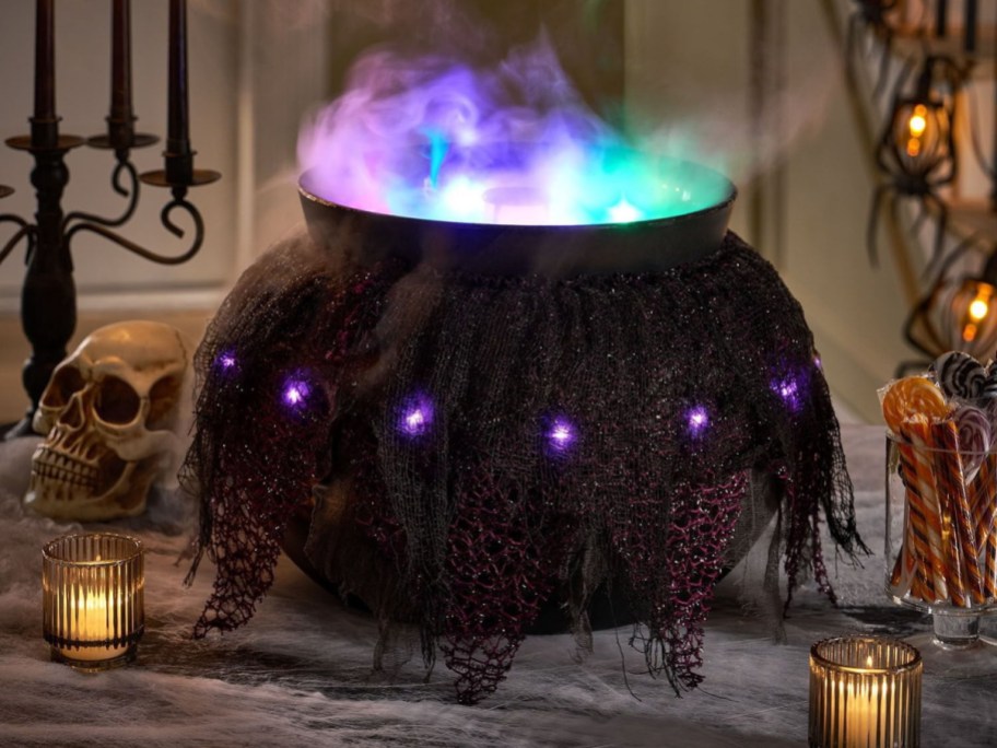 A lighted Halloween cauldron decoration draped in dark tattered fabric with embedded purple lights, emitting fog and glowing multi-colored lights from within, displayed on a table alongside a skull, lit candles, and candy.