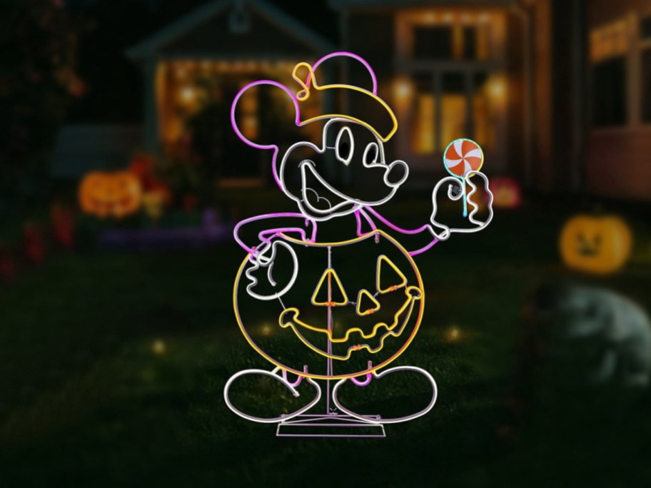A neon-style light-up yard decoration of Mickey Mouse standing inside a jack-o'-lantern holding a lollipop, set on a dark lawn at night.