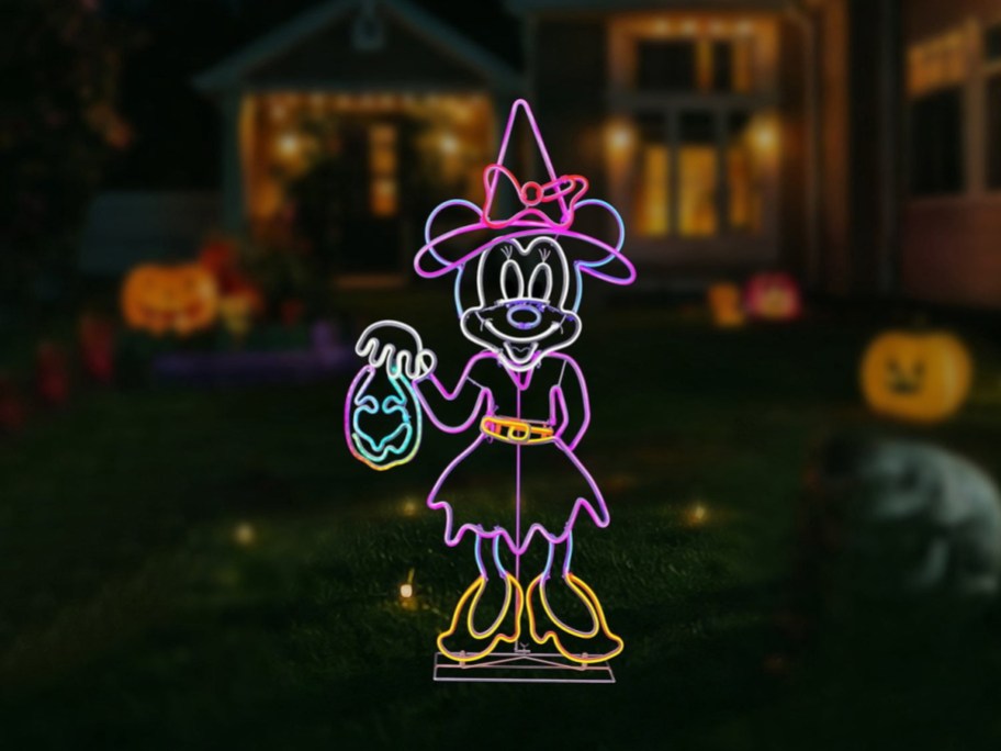 A neon-style light-up yard decoration of Minnie Mouse dressed as a witch holding a trick-or-treat bag, set on a dark lawn at night.