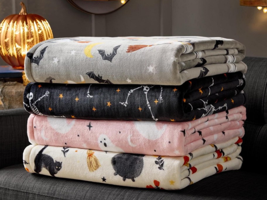 A stack of four folded Halloween-themed plush throw blankets resting on a dark sofa, featuring patterns of bats, skeletons, ghosts, and black cats, with a lit glass pumpkin in the background.