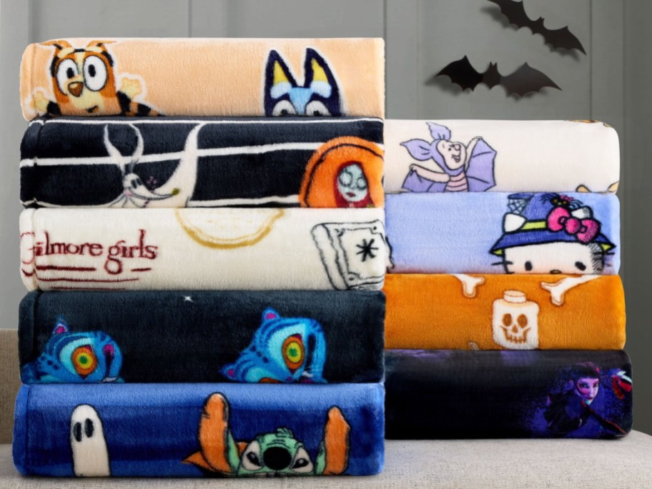 Two stacks of folded plush throw blankets featuring various pop culture and Halloween character designs, displayed against a grey wall with black bat silhouettes.