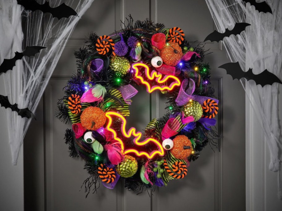 A brightly lit Halloween wreath hanging on a grey door, decorated with glowing neon bats, glittery pumpkins, eyeballs, colorful mesh, and string lights, surrounded by fake cobwebs and black bat silhouettes.
