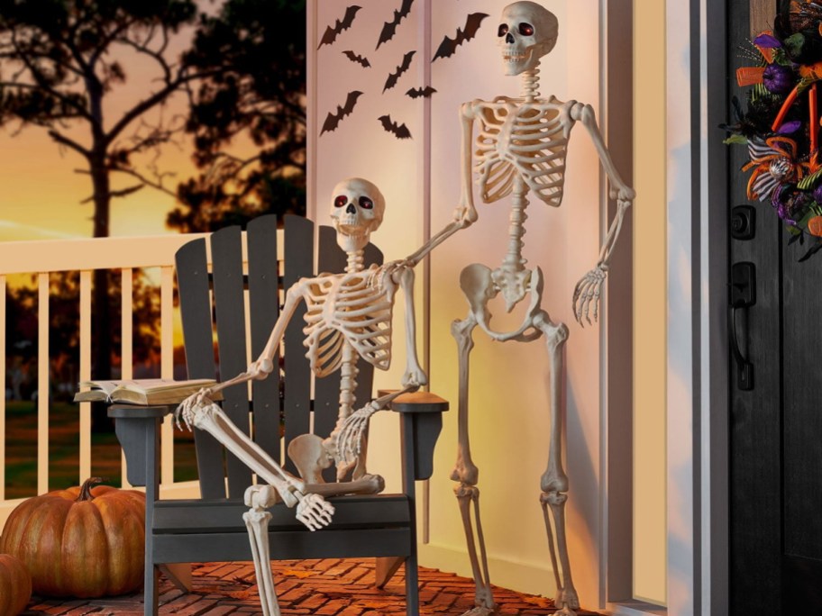 Two Halloween skeletons with glowing red eyes on a porch at sunset, one sitting in an Adirondack chair and the other standing, decorated with pumpkins and bat silhouettes.
