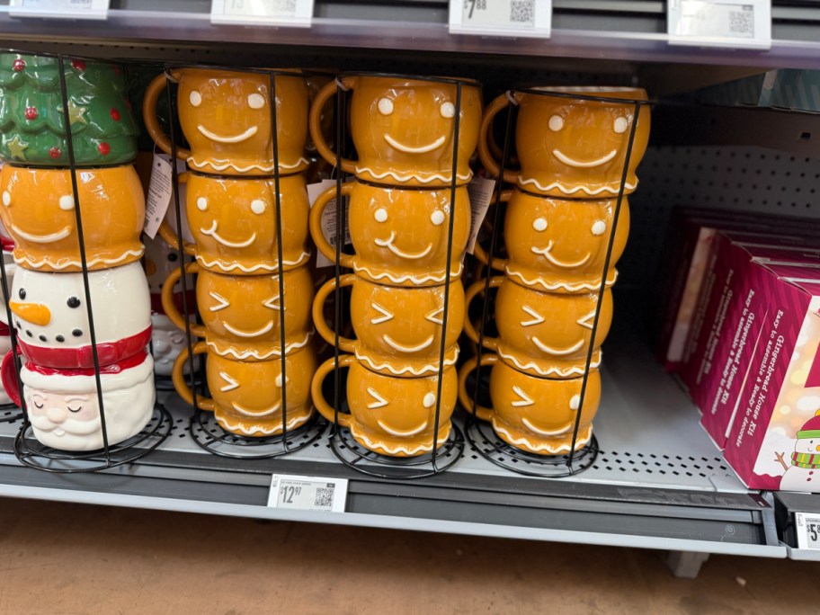 Multiple vertical racks of stacked holiday mugs on a store shelf, including stacks of yellow gingerbread person mugs and a stack of assorted holiday character mugs.