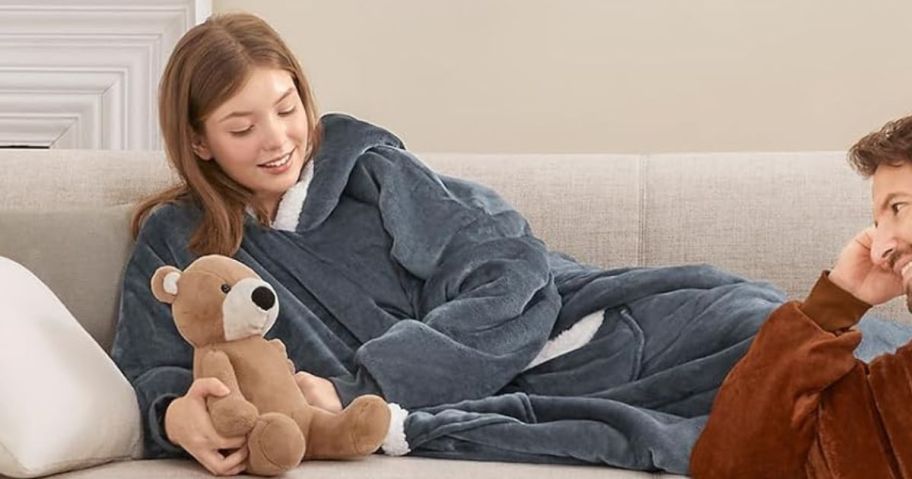 woman laying on couch while wearing grey wearable blanket hoodie and holding teddy bear