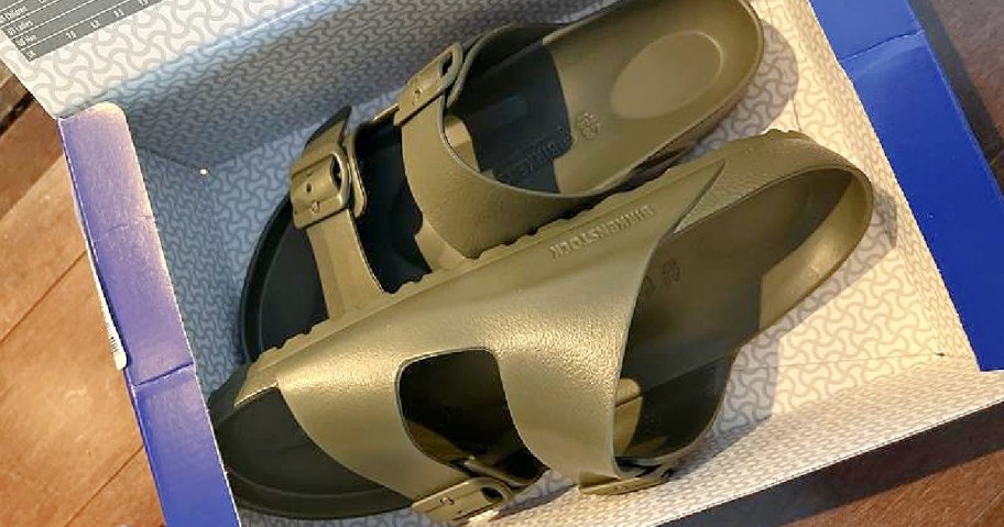 A pair of olive green sandals with buckles, placed in an open blue shoebox on a wooden floor. The sandals appear new and neatly arranged.