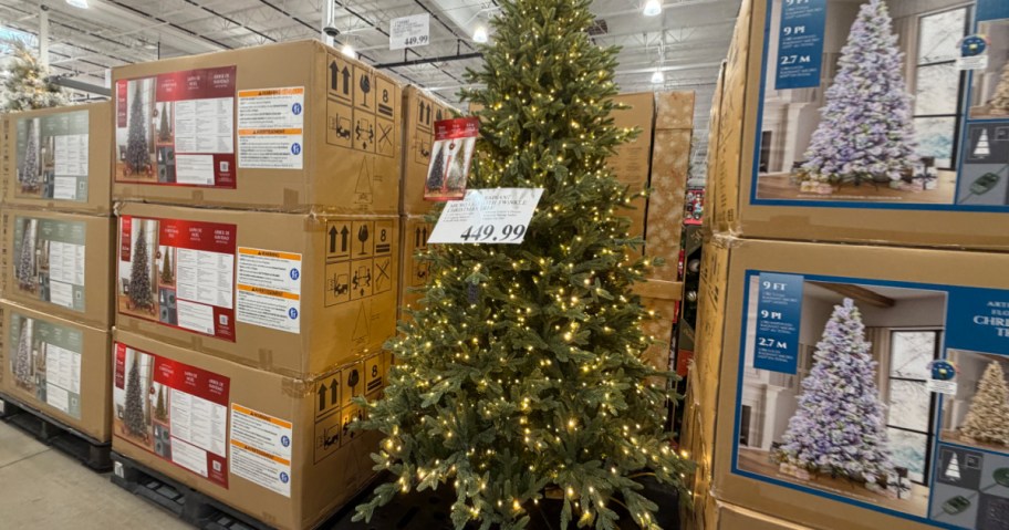 A pre-lit Christmas tree displayed on a store floor, surrounded by stacks of boxed trees.