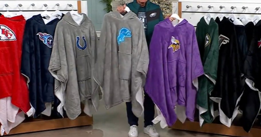 man holding 2 NFL wearable blanket hoodies, more on display racks on each side of him