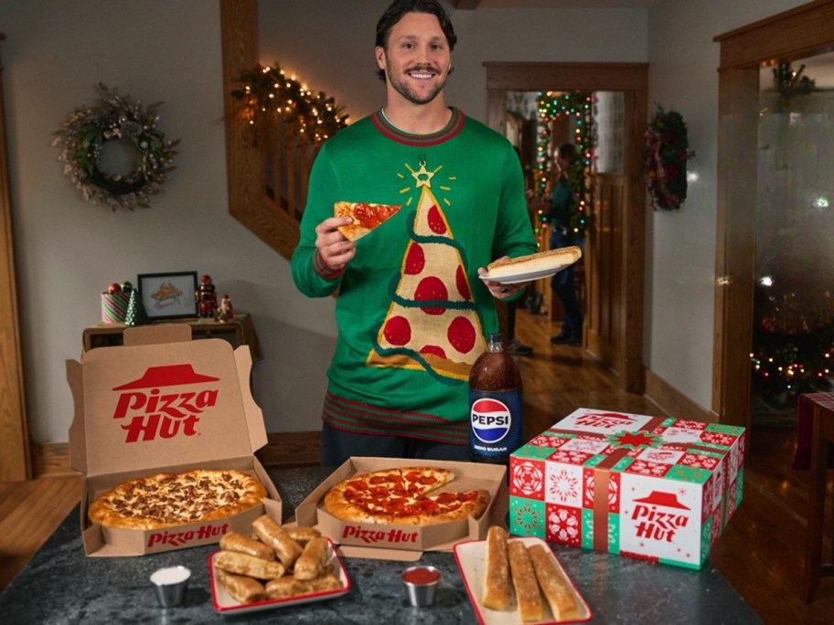 man wearing a sweater with a pizza slice on it, holding a pizza slice, Pizza Hut boxes and food on a table in front ofhim