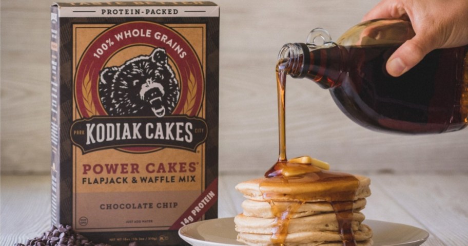 box of kodiak cakes chocolate chip pancake and waffle mix behind stack of pancakes with syrup being poured over them