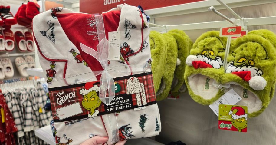 A person holding a pair of Grinch pajamas up in front of a pair of Grinch slippers in a store