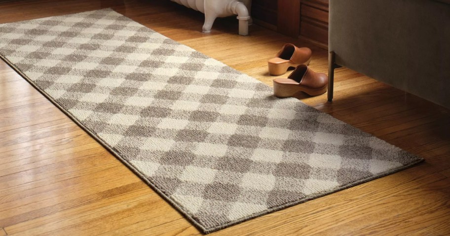A plaid wool runner with a neutral checkered pattern lies on a wooden floor beside a couch. Two brown clogs are placed neatly at its end.