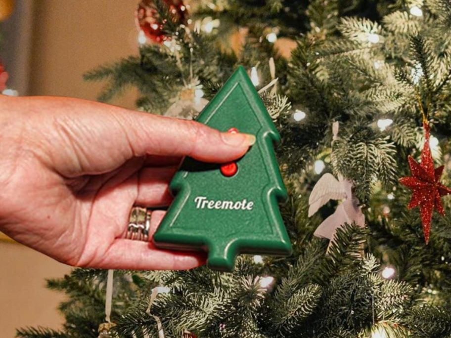 A person holding a remote shaped like a tree in front of a lit Christmas Tree