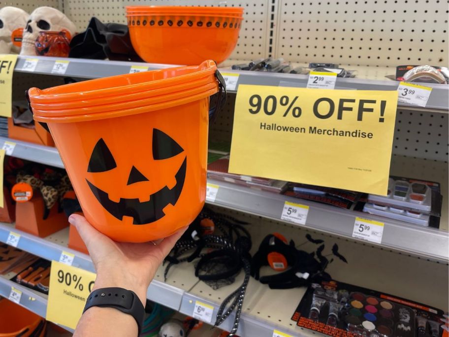 A person holding a jack-o-lantern bucket next to a 90% off sign