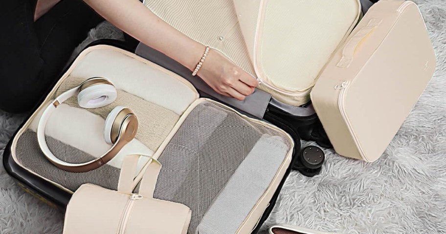 A person packs a suitcase with beige packing cubes, headphones, and folded clothes. Everything rests on a plush gray carpet.