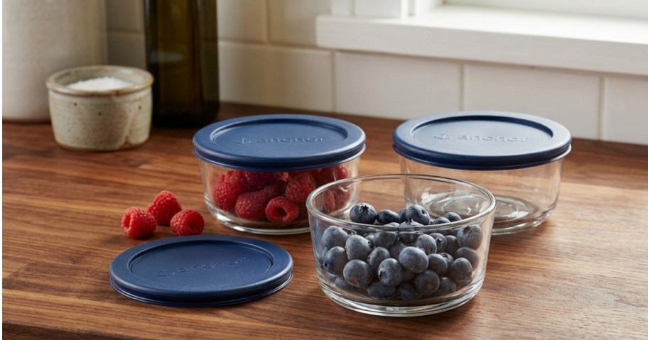 Glass bowls filled with raspberries and blueberries are on a wooden countertop, each with a blue lid nearby.