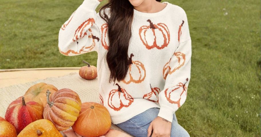 woman wearing bloomchic sweater with pumpkins on it sitting next to pile of pumpkins