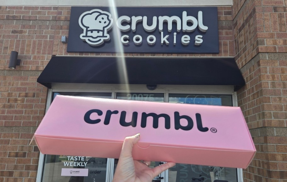 womans hand holding a pink crumble box up infront of a crumbl store front