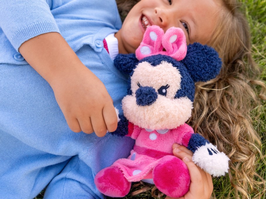 A joyful child in a blue sweater lies on grass, holding a pink-dressed plush toy of Minnie Mouse.