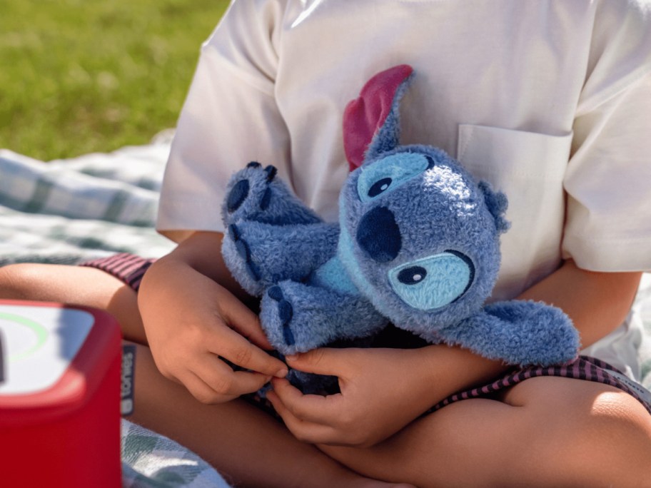 Child holding a blue plush Stitch toy with big eyes, sitting cross-legged on a picnic blanket outdoors.