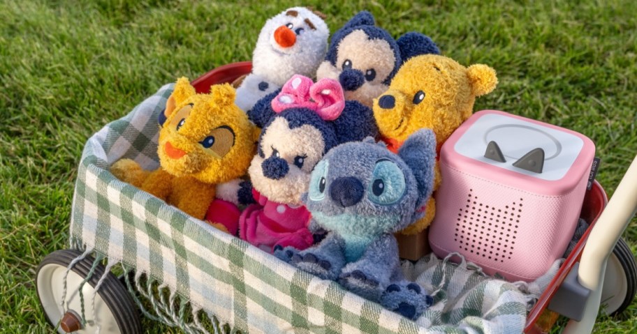 A group of colorful plush toys is nestled in a green and white wagon on a grassy lawn. A pink speaker sits beside them.