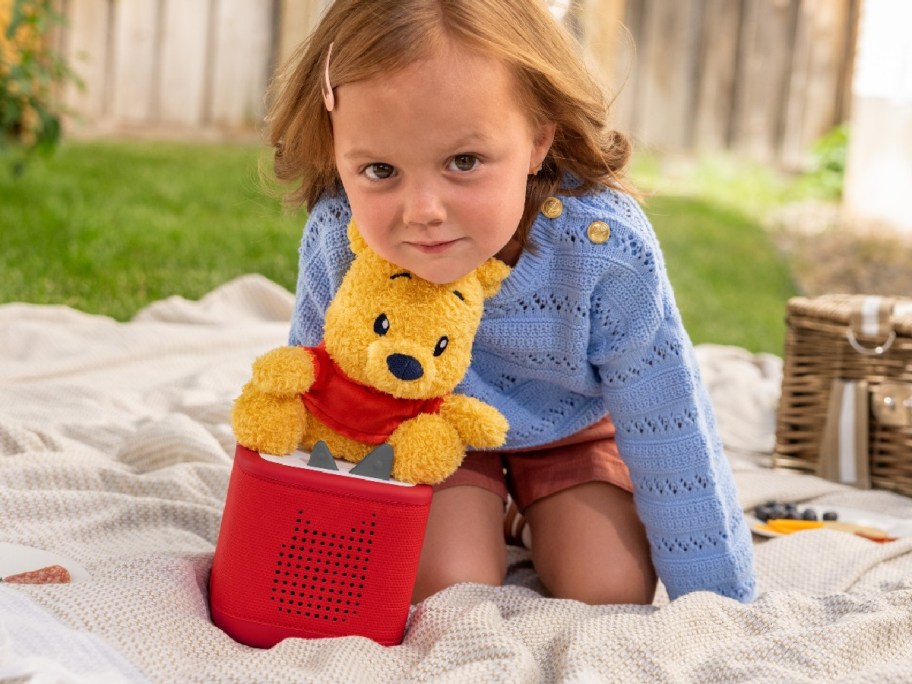young girl with a plush winnie the pooh toy sitting on top of a tonie box
