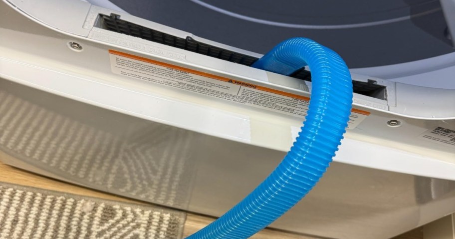 A close-up of a dryer's lint trap with a bright blue hose sticking out. Textured gray mat partially visible on wooden floor.