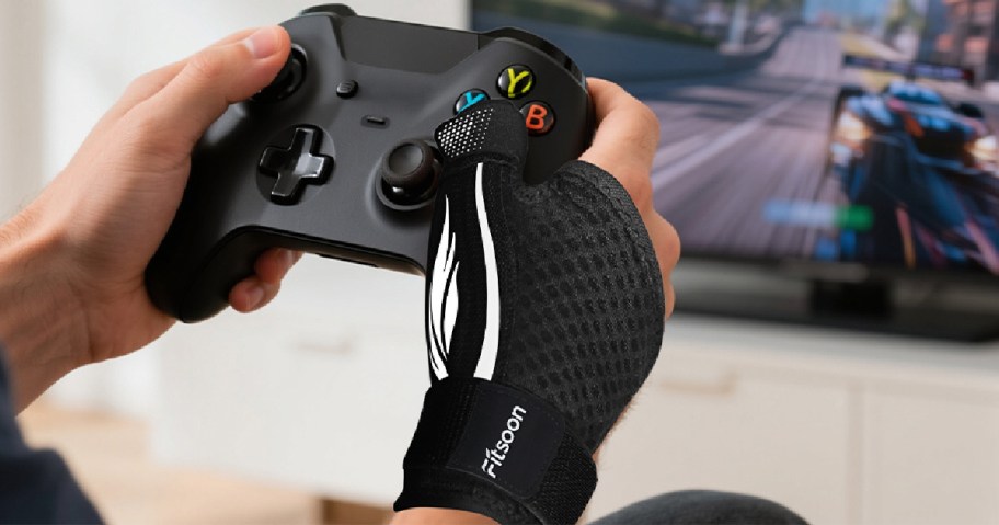 Hands are holding a black gaming controller, with one hand wearing a black wrist support. A racing game is displayed on a TV in the background.