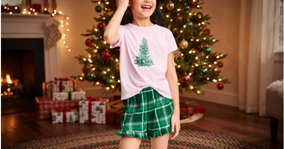 Cat & Jack girls' holiday pajama set featuring a light pink short-sleeve shirt with a decorated Christmas tree graphic and green plaid ruffle shorts, standing in front of a lit Christmas tree and fireplace.
