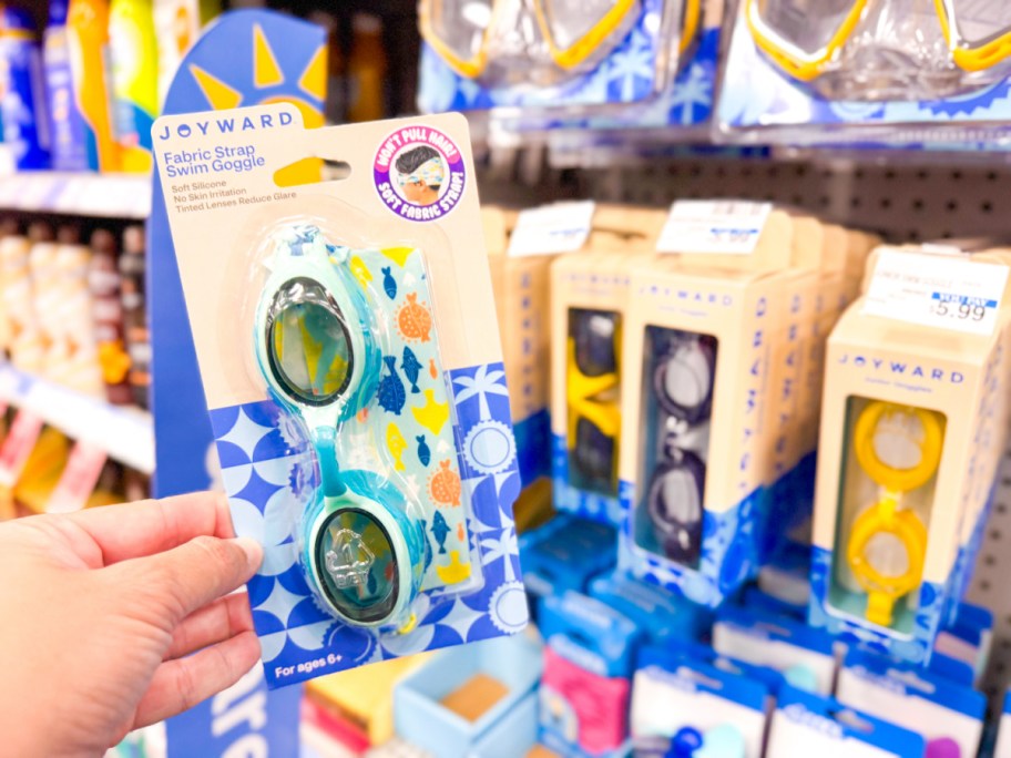 A hand holding a package of light blue Joyward swim goggles in a store aisle.