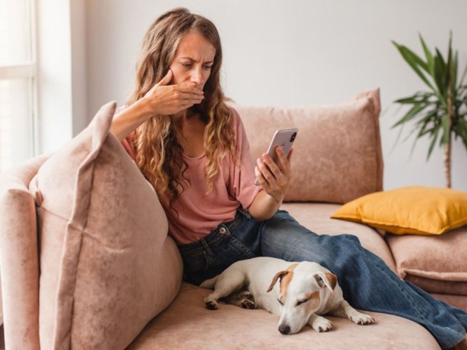 A woman holding a mobile phone and putting her hand over her mouth while sitting on the sofa