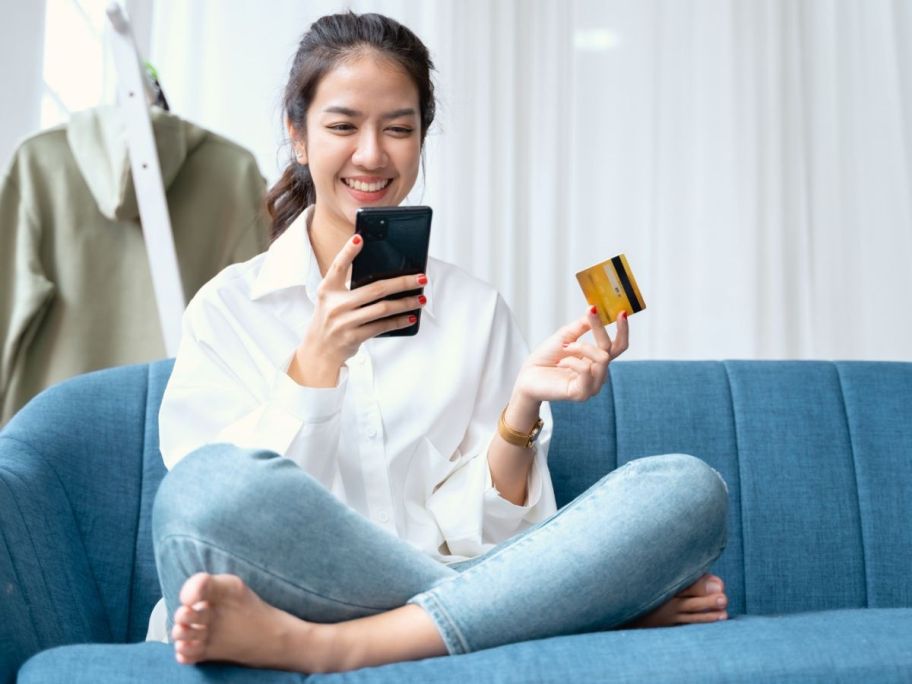 A woman sitting on the sofa smiling and looking at her mobile phone while holding a credit card
