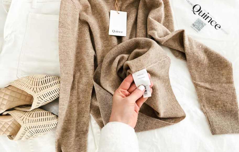 hand holding a quince tag on brown sweater laying on bed with sandals