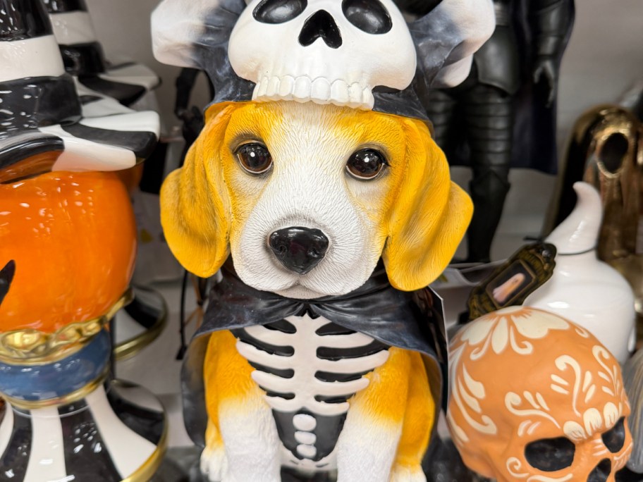 A figurine of a beagle puppy wearing a skeleton costume and a skull hat, surrounded by other Halloween decor items.